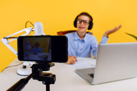 student is studying at home on distance learning, sitting at a Desk and shooting an online broadcast from a mobile phone on a tripod, on a yellow backgroundの写真素材