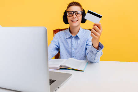 schoolboy guy is sitting at a table with a laptop and holding a credit card on a yellow background....