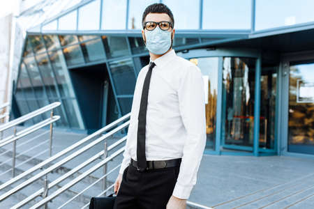 young businessman in business clothes, a man in a medical protective mask on his face, leaves the office building after a working dayの写真素材