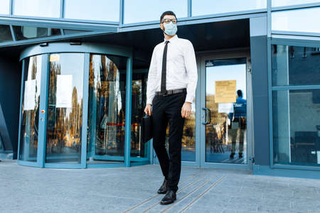 Man businessman in business clothes and a medical protective mask on his face, against the background of a glass office buildingの写真素材