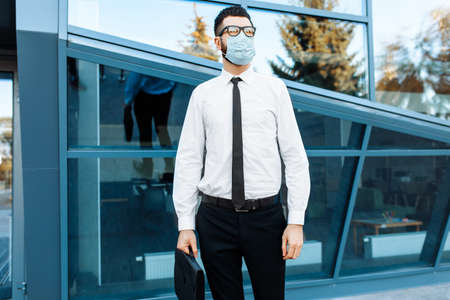 Man businessman in business clothes and a medical protective mask on his face, against the background of a glass office buildingの写真素材