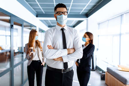 Confident young business man leader wearing a medical protective mask on his face, with a group of colleagues in the background, standing in an office buildingの写真素材