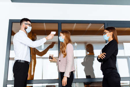 man with a non-contact thermometer measures the temperature of employees in protective masks at the entrance to a modern officeの写真素材