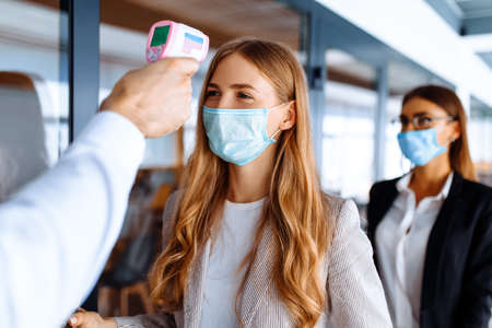 man with a non-contact thermometer measures the temperature of employees in protective masks at the...