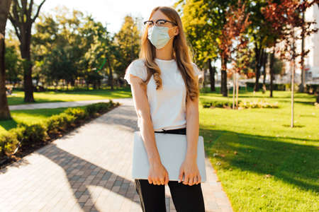 Young attractive woman in a medical protective mask on her face, with a laptop in the city concept of coronavirusの写真素材