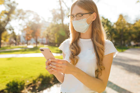 Young business woman in a medical protective mask on her face uses a mobile phone on the street in the cityの写真素材