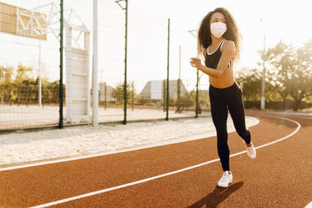 Beautiful african american woman in medical protective mask on her face, jogging outdoors, fitness,...