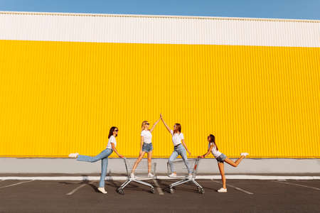Group of happy young people having fun on shopping carts. Young beautiful girlfriends rush on a cart on a sunny summer day against the background of a yellow wall of a shopping centerの写真素材