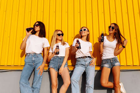 Attractive happy female friends in casual clothes posing and having fun and drinking drinks on the background of the yellow wall of the shopping mall on a sunny summer summer dayの写真素材