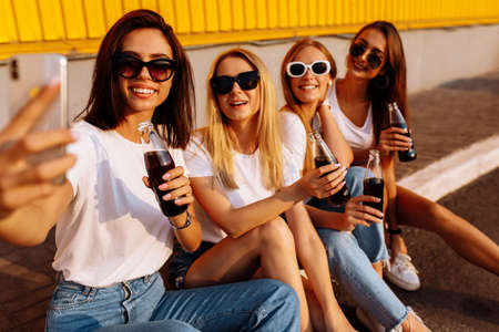 Young friends drinking a drink having fun together and taking a selfie on a mobile phone, against the background of a yellow wall, the concept of friendship and celebrationの写真素材