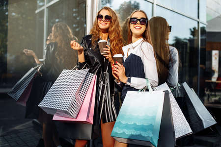 Two happy beautiful female friends in sunglasses, with shopping bags, drink coffee in paper cups, near a shopping centerの写真素材