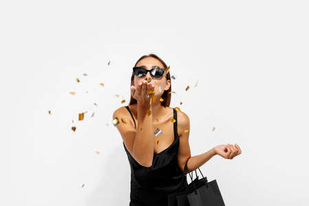 Excited young woman, in black dress, with shopping bags, celebrating and throwing confetti on white background, concept, shopping, Black Fridayの写真素材