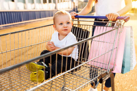 Excited young mother with baby and shopping bags and shopping trolley near mall