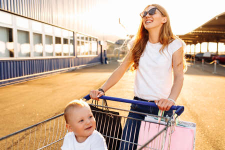 Happy young mother with baby and shopping bags and shopping trolley near mall