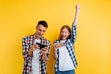 happy couple, a man and a woman, play video games on a smartphone, rejoice and show a winning gesture, yellow backgroundの写真素材