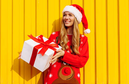 Attractive happy young woman in christmas sweater and santa claus hat with christmas present...