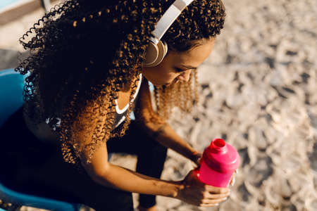 Young athletic african american woman wearing headphones resting after intense jogging and drinking water from a bottle during a break after jogging outdoorsの写真素材
