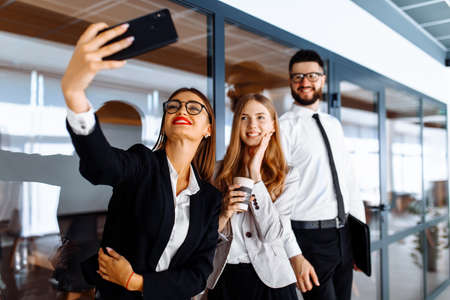 Group of attractive business people standing and chatting together holding coffee cups and taking selfie on mobile phone in modern officeの写真素材