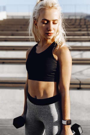 Athletic young woman working out with dumbbells, doing exercises for the arms, outdoors