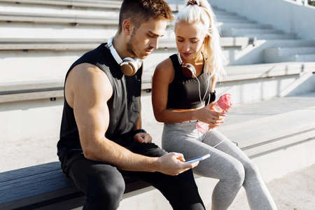 Young sports couple, in sportswear, sitting on the stairs outdoors, listening to music on headphones...