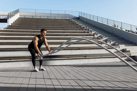 Strong man exercising with crossfit fighting ropes, muscular athlete doing cross excursion with...