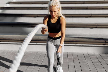 Sporty young woman, in sportswear, doing crossfit exercises with fighting ropes, outdoors in the...