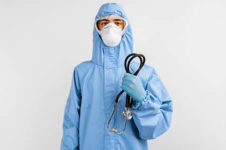doctor in a blue protective suit, mask, glasses and gloves holds a stethoscope on a white background, the threat of a coronavirus pandemicの写真素材