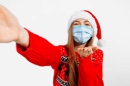Happy attractive young woman, in a Christmas sweater and Santa Claus hat, wears a medical protective mask from viruses, makes a selfie in the studio on a white background, New Year, Christmasの写真素材