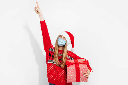 Beautiful excited young woman wearing Christmas Santa Claus hat and medical mask on her face holding...