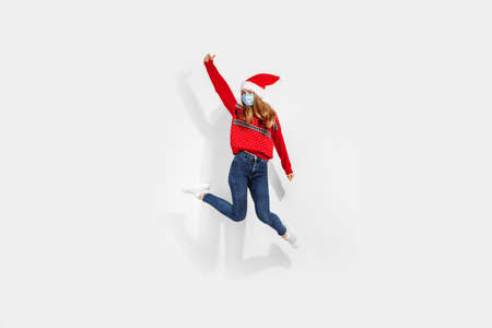 Full length of an excited cheerful young Christmas woman wearing a red Santa Claus hat and a medical protective mask on her face, having fun and jumping against a white background,の写真素材