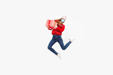 Full length of an excited cheerful young Christmas woman wearing a red Santa Claus hat and a medical protective mask on her face, having fun and jumping with a gift box against a white background,の写真素材