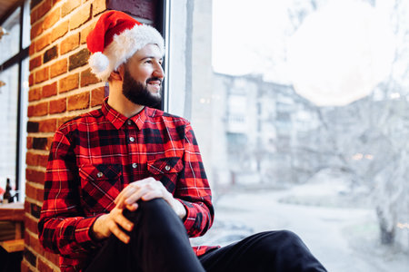 Attractive christmas man wearing santa claus hat sits on windowsill and looks out the window enjoying christmas in stylish homeの写真素材