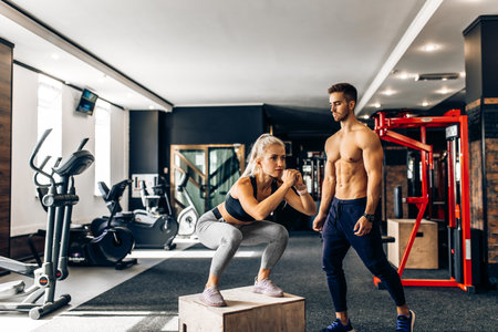 Strong young woman jumping on Fit Box exercising with box in the gym with male trainer, athlete doing workout in the gym with instructorの写真素材