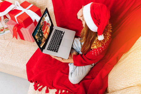Happy little girl wearing Santa Claus hat, talking to parents by video link, celebrating Christmas and New Year, talking at a remote online conference on a computer at home.の写真素材