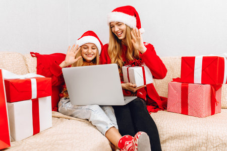 Happy family, mom and little daughter in santa claus hats are holding a Christmas video call with their family, video conference on skype, waving their hands, congratulating relatives sitting on the couch at homeの写真素材