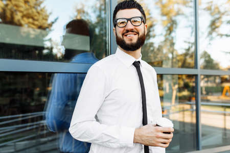 Young bearded businessman in glasses walking down a city street holding a cup of coffee, against the background of a modern glass building, a man rushing to a business meetingの写真素材