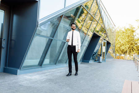 Successful business man with laptop holding a cup of hot drink in hand on his way to work on city...