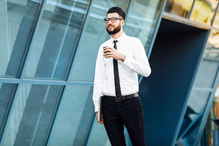 Successful business man with laptop holding a cup of hot drink in hand on his way to work on city...