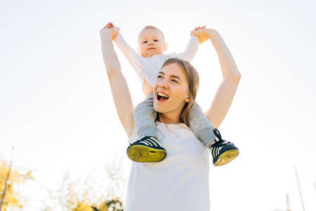 Happy young mother around her neck with her baby playing and having fun with her little son, on a sunny dayの写真素材
