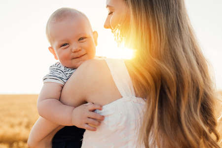 Happy family, beautiful young mother in a dress with a cute little baby on a golden wheat field at sunset on a summer dayの写真素材