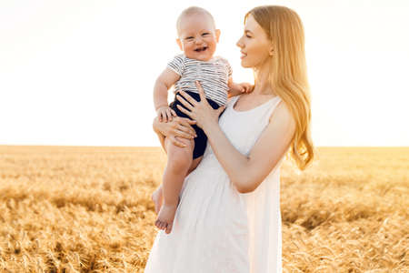 Happy beautiful mother with her baby on the golden wheat field, family on the grain field, mom hugs her little boyの写真素材