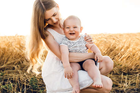 Happy beautiful mother with her baby on the golden wheat field, family on the grain field, mom hugs her little boyの写真素材