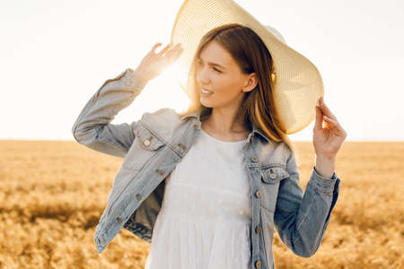 Happy beautiful young woman in dress and hat in a wheat field at sunsetの写真素材