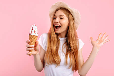 Happy beautiful young woman, in a summer hat, with a huge ice cream cone on a pink backgroundの写真素材