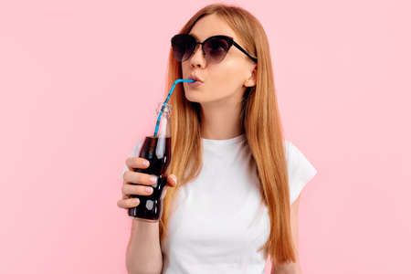 Beautiful young woman in sunglasses, in a white t-shirt, drinks a refreshing drink from a glass bottle on an isolated pink backgroundの写真素材