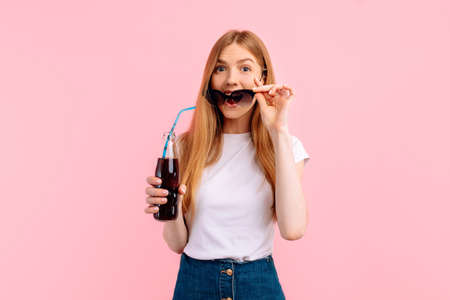Portrait of excited young woman wearing sunglasses holding soda on pink backgroundの写真素材