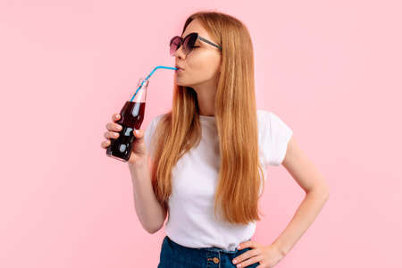 Portrait of cute beautiful young woman in sunglasses drinking soda from bottle, on isolated pink backgroundの写真素材