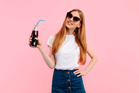 Happy young woman wearing sunglasses holding soda bottle isolated on pink backgroundの写真素材