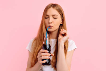 Portrait of cute beautiful young woman in sunglasses drinking soda from bottle, on isolated pink backgroundの写真素材