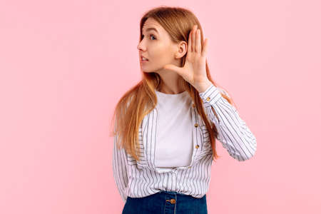 Curious young woman holding hand to ear trying to eavesdrop on conversation, on pink studio backgroundの写真素材